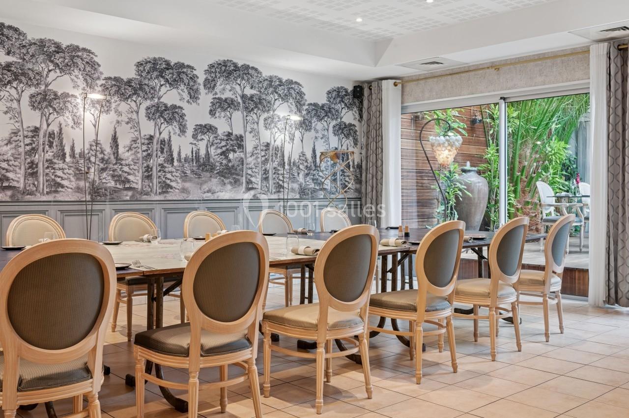Salle à manger lumineuse avec une grande table entourée de chaises, décorée d'une fresque murale et ouverte sur un jardin.