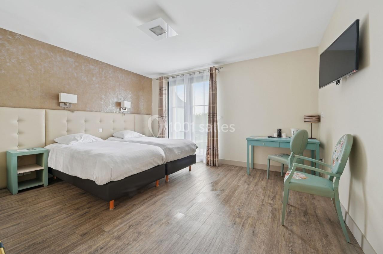 Chambre lumineuse avec deux lits simples, sol en bois, bureau, chaise, télévision murale et grande fenêtre avec rideaux.