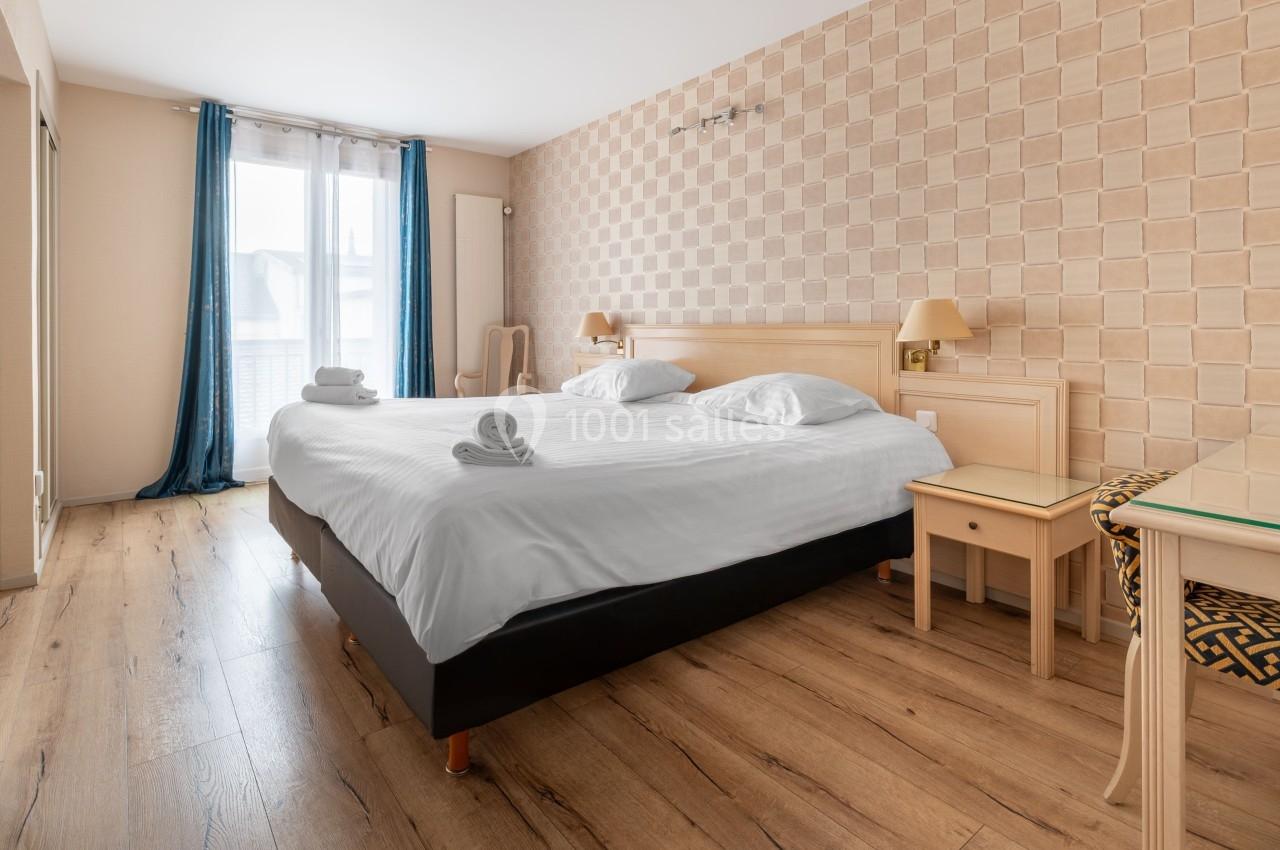 Chambre lumineuse avec un grand lit, parquet au sol, murs beige texturés et rideaux bleus encadrant une fenêtre.