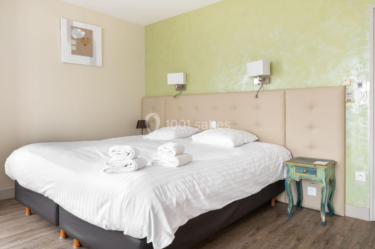 Lit double avec tête de lit beige, draps blancs, serviettes pliées, mur vert clair et table de chevet vintage.