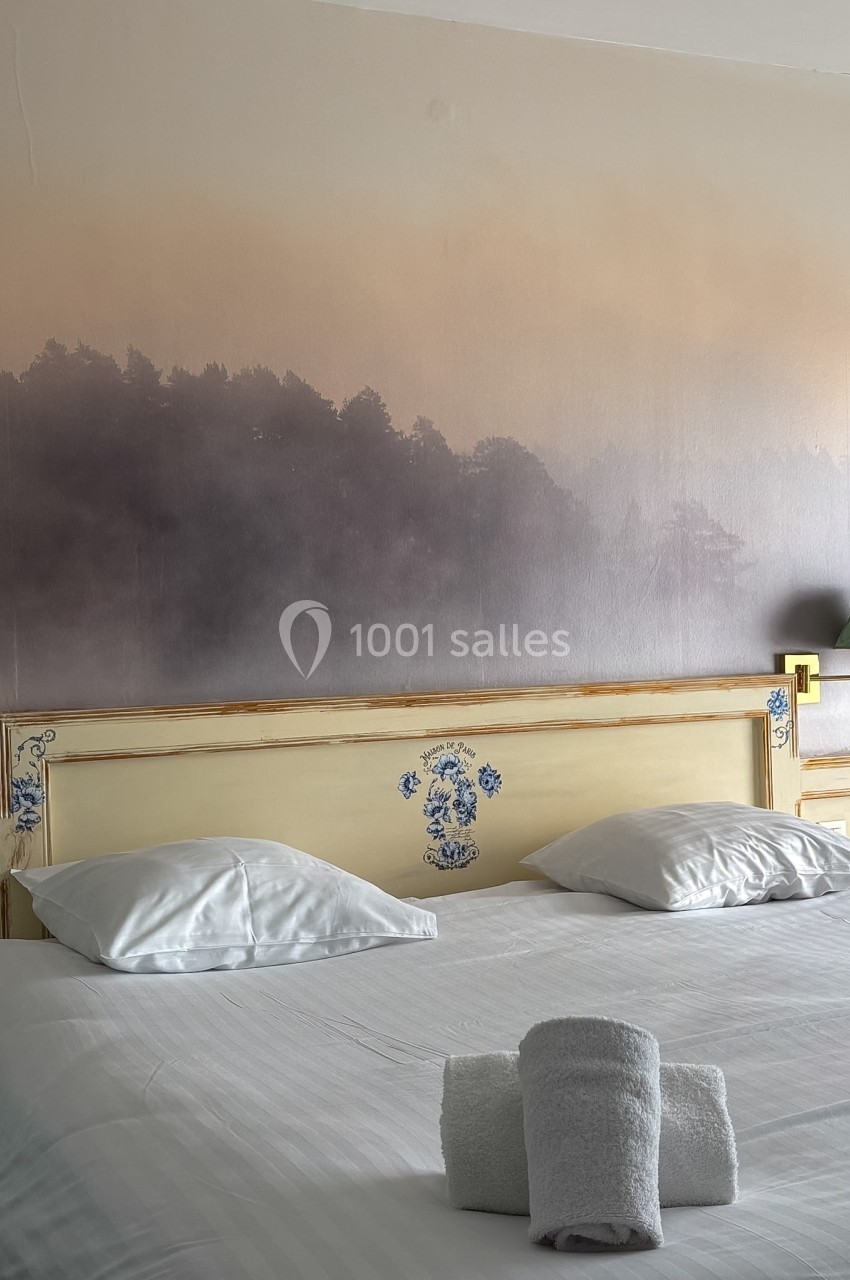 Lit double avec tête de lit décorée, draps blancs et serviettes pliées, devant un mur avec un motif de forêt brumeuse.