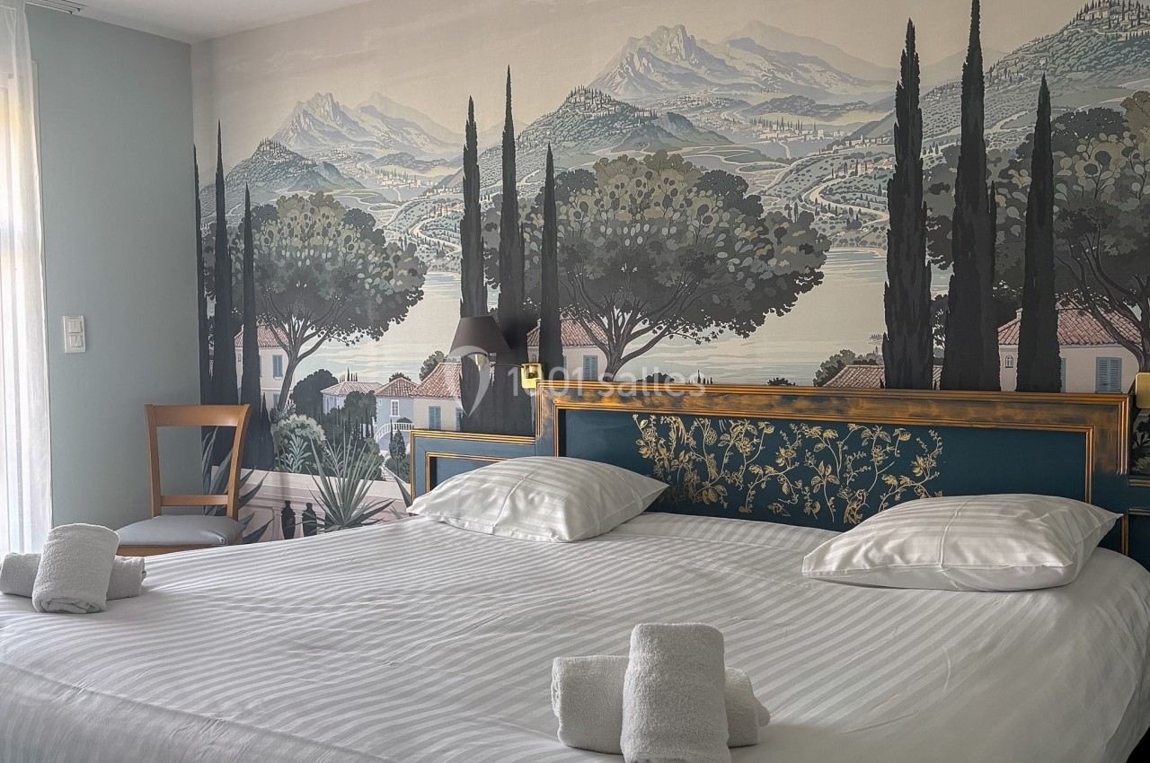 Lit double avec tête de lit décorée, draps blancs et serviettes pliées, devant une fresque murale de paysage méditerranéen.
