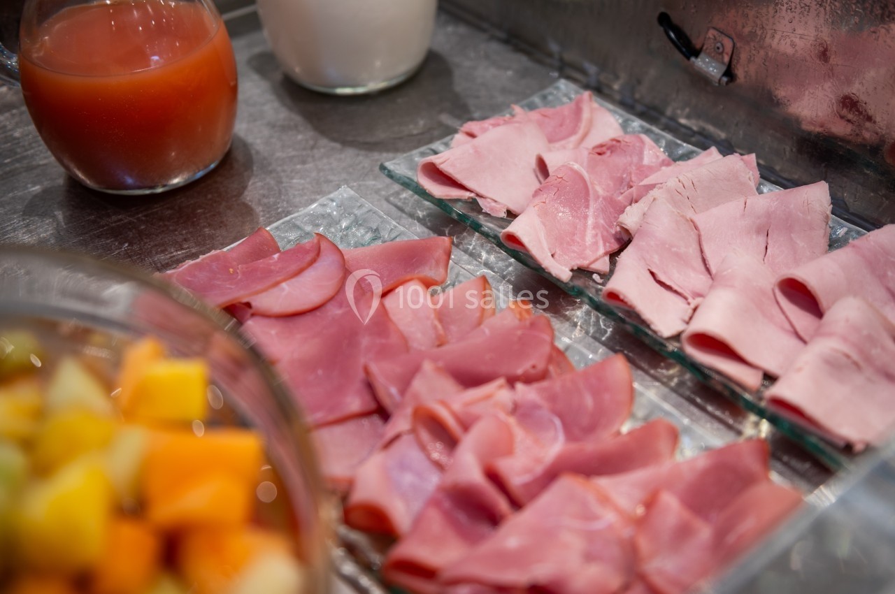 Tranches de jambon rose disposées sur des assiettes en verre, avec fruits et jus en arrière-plan.