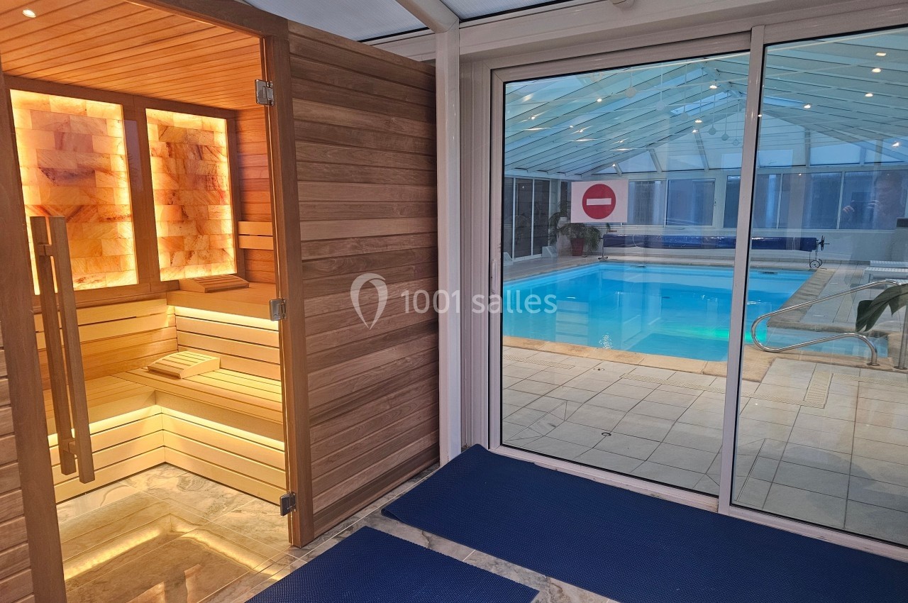 Vue d'un sauna en bois éclairé à gauche et d'une piscine intérieure derrière une baie vitrée à droite.