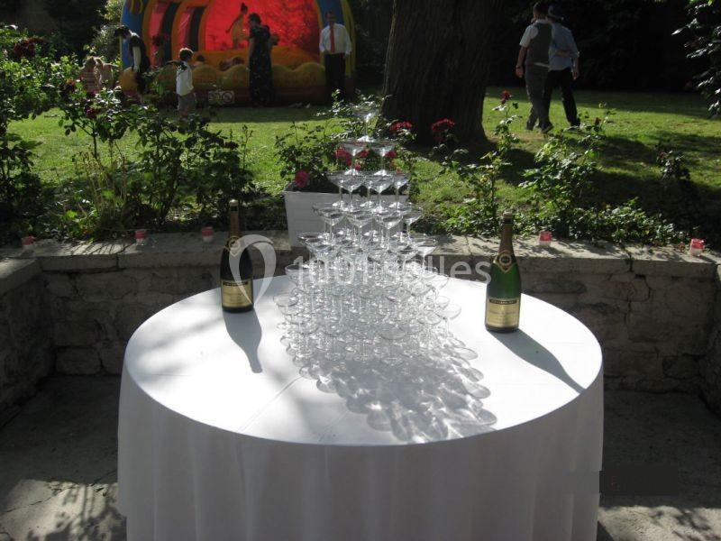 Pyramide de coupes à champagne sur une table blanche, entourée de bouteilles et située dans un jardin ensoleillé.