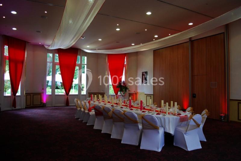 Salle de réception décorée avec des nappes blanches, des chaises ornées de rubans dorés et des rideaux rouges.