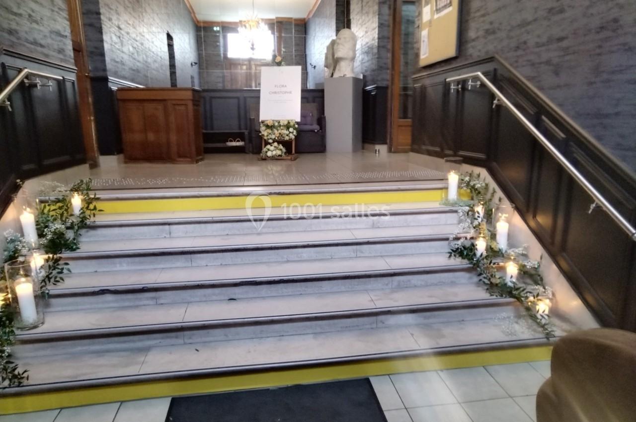 Escalier décoré de bougies et de fleurs blanches, menant à une salle avec une statue et un panneau d'information.