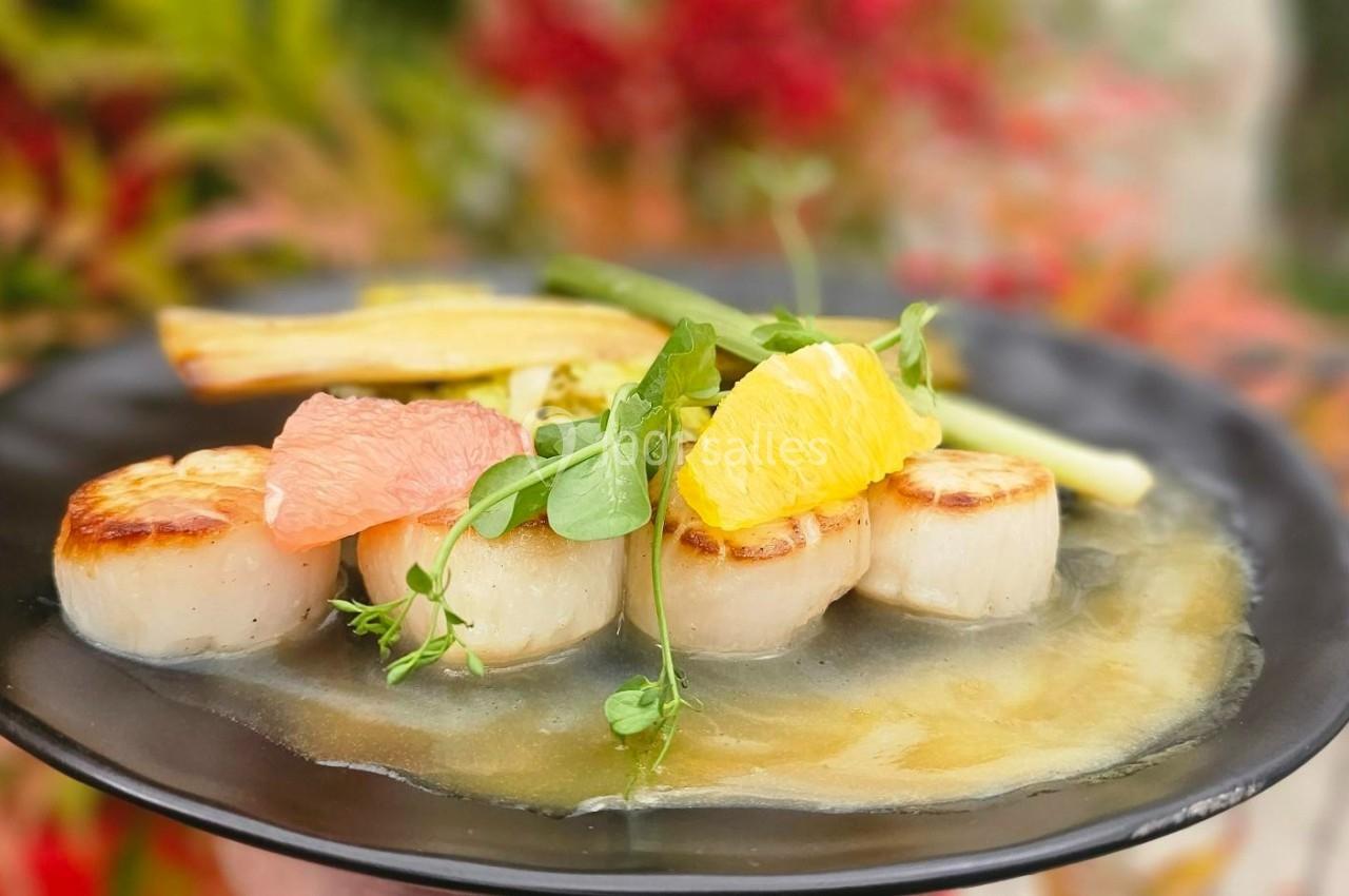 Assiette de noix de Saint-Jacques poêlées, accompagnées de légumes, agrumes et herbes fraîches sur un fond flou.