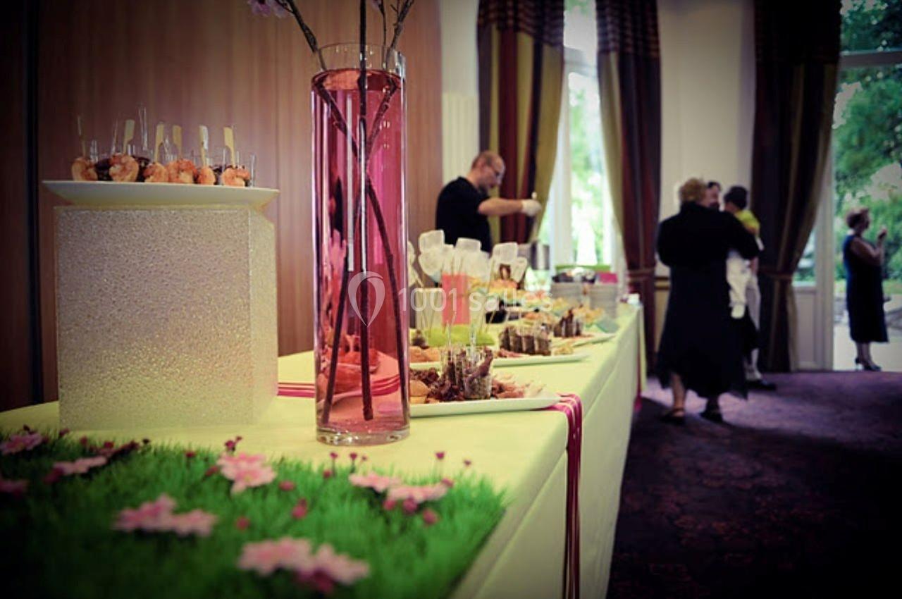 Buffet décoré avec des fleurs, des verrines et des plats variés, dans une salle avec des rideaux et des invités en arrière…