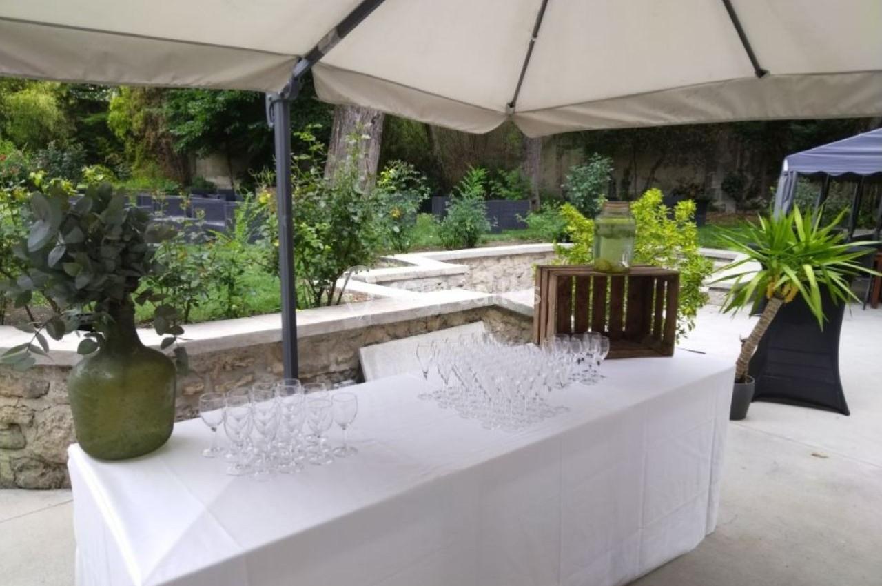 Table dressée en extérieur avec des verres, un vase vert et une caisse en bois sous un grand parasol.