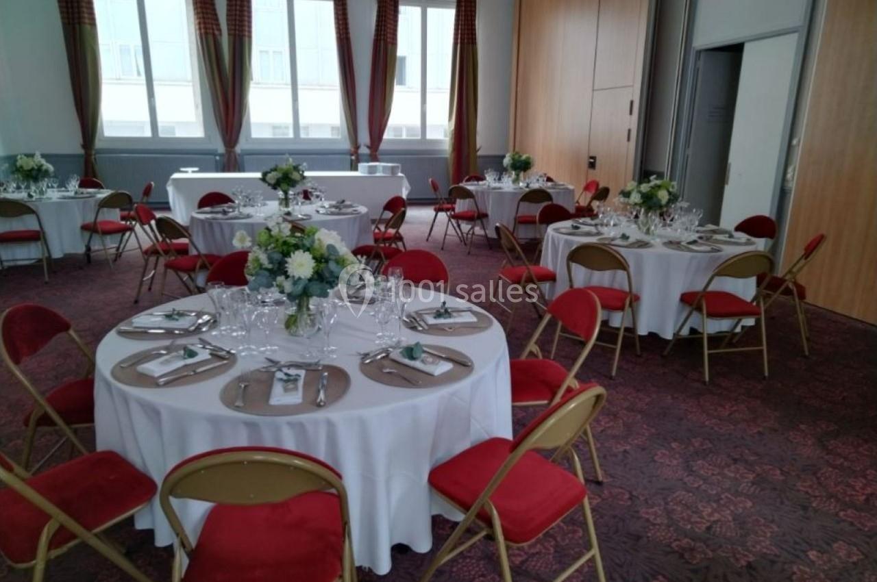 Salle de réception avec tables rondes dressées, nappes blanches, chaises rouges et décorations florales.