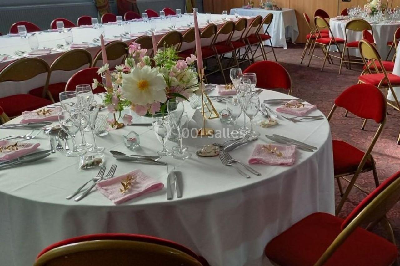Salle de réception avec tables rondes dressées, nappes blanches, chaises rouges et décoration florale au centre.