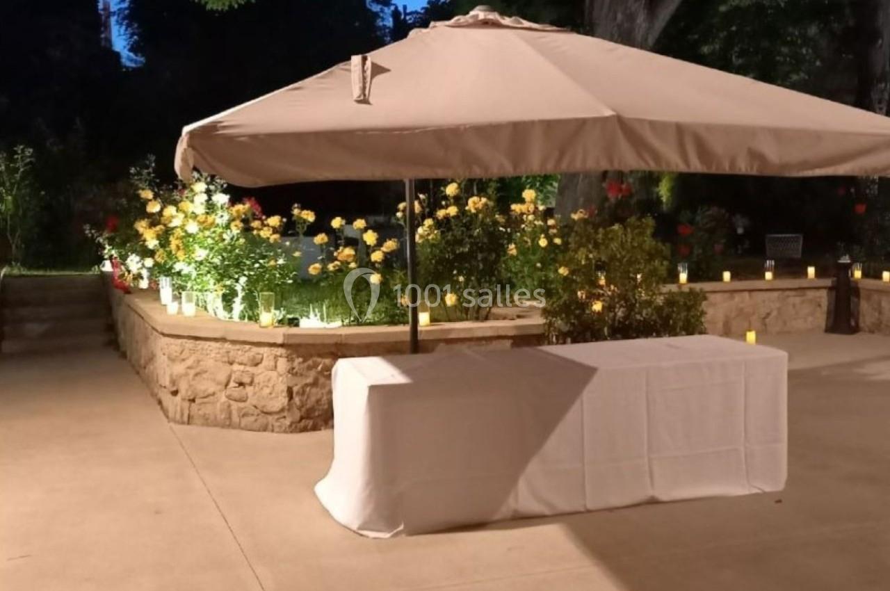 Parasol beige et table recouverte d'une nappe blanche dans un jardin éclairé en soirée, entouré de fleurs et de bougies.