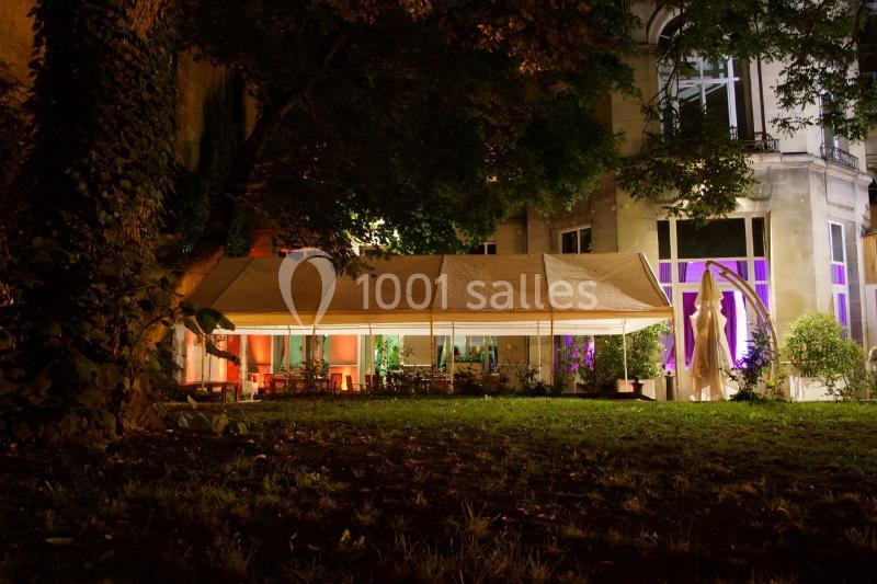 Tente éclairée installée dans un jardin, entourée d'arbres et d'un bâtiment aux fenêtres illuminées.