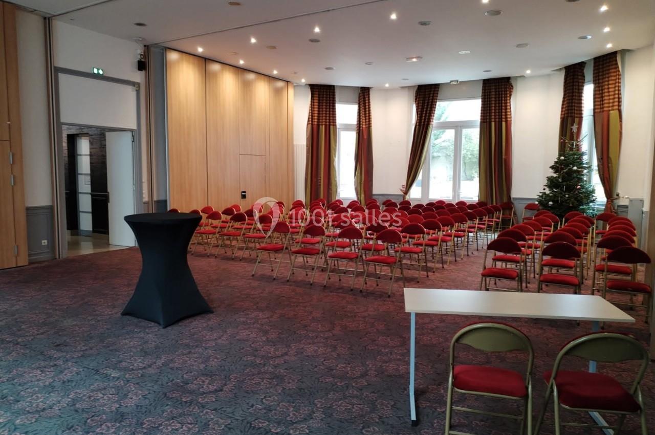 Salle de conférence avec des rangées de chaises rouges, une table blanche et un pupitre noir, éclairée par des fenêtres.