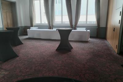 Salle vide avec moquette, tables hautes noires et une longue table blanche près des fenêtres avec rideaux.