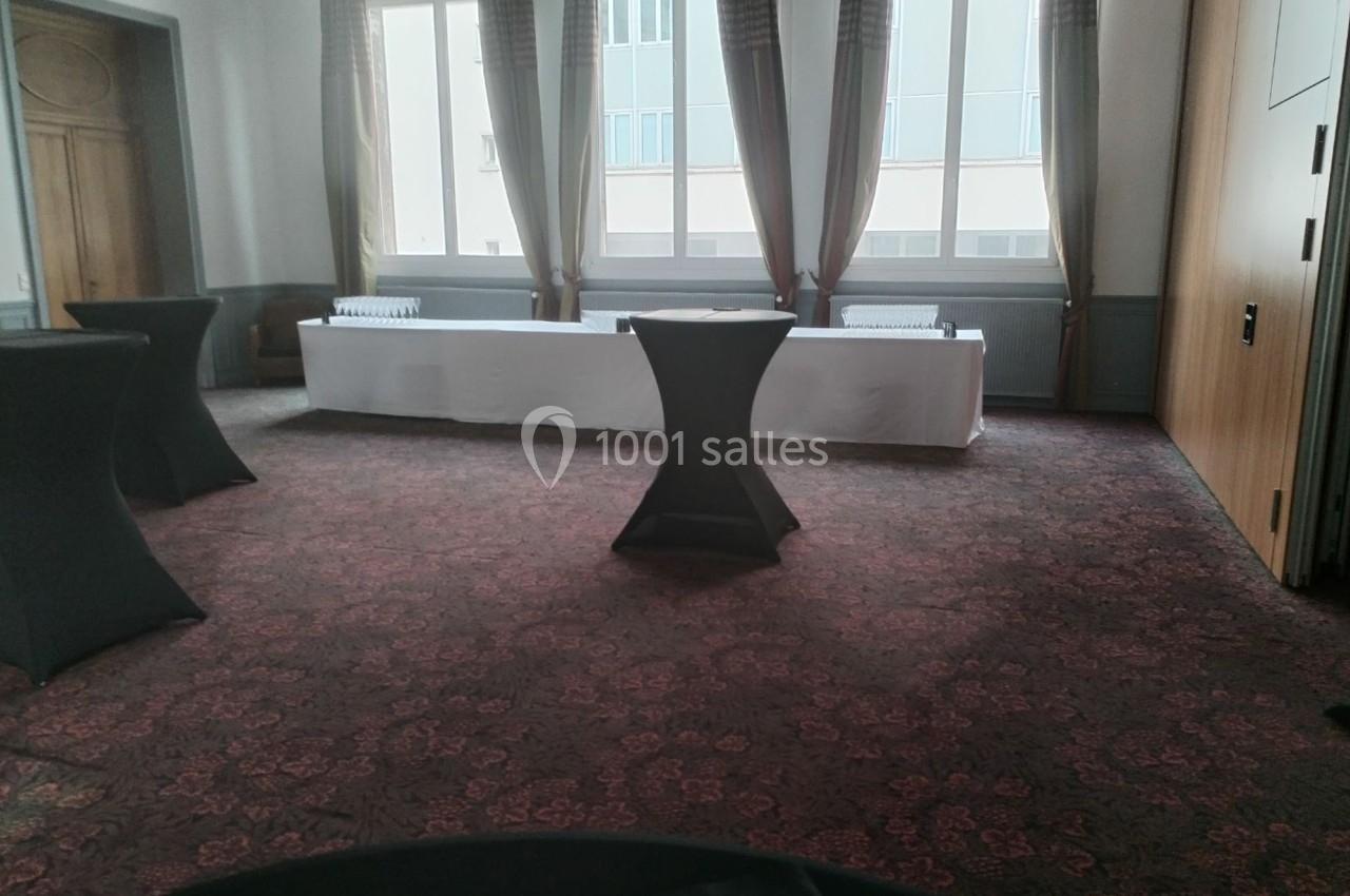 Salle vide avec moquette, tables hautes noires et une longue table blanche près des fenêtres avec rideaux.