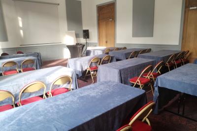 Salle vide avec moquette, tables hautes noires et une longue table blanche près des fenêtres avec rideaux.