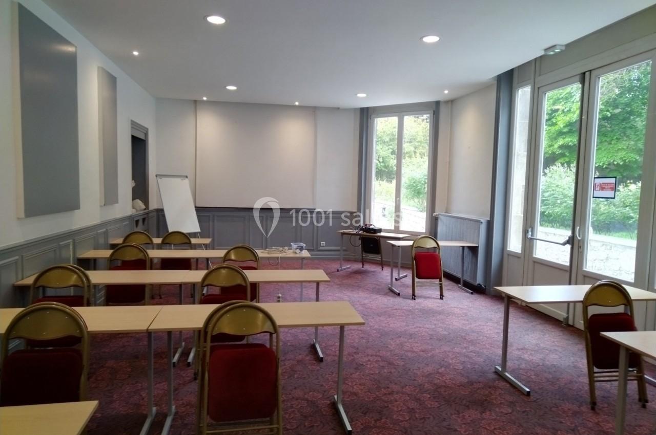 Salle de réunion lumineuse avec des tables, des chaises, un tableau blanc et de grandes fenêtres donnant sur un espace vert.
