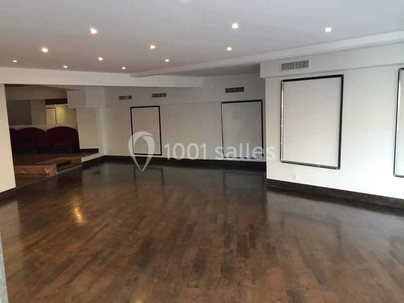 Salle vide avec parquet en bois sombre, murs blancs, cadres vides et éclairage par spots encastrés au plafond.