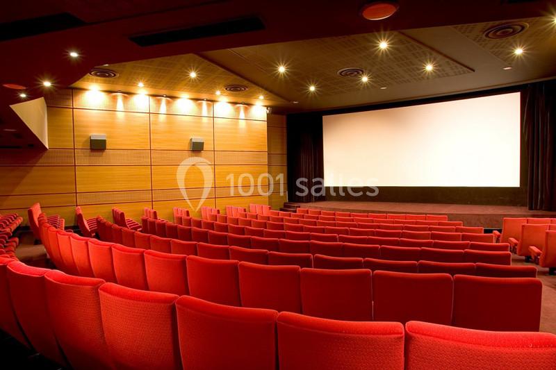 Salle de cinéma vide avec des rangées de sièges rouges, un écran blanc et un éclairage tamisé au plafond.