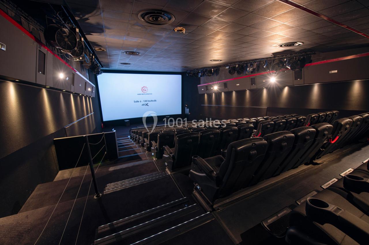 Salle de cinéma vide avec rangées de sièges noirs, écran blanc allumé et éclairage tamisé.