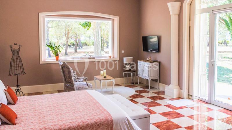 Chambre lumineuse avec lit double, fauteuil près d'une grande fenêtre, sol en carreaux rouges et blancs, et vue sur jardin.