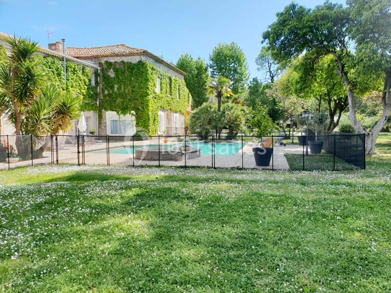 Maison en pierre recouverte de lierre, avec piscine clôturée et jardin arboré sous un ciel bleu.