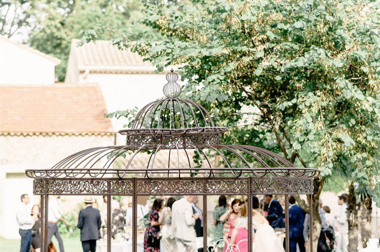Structure en fer forgé dans un jardin, avec des invités rassemblés en arrière-plan près d'un arbre.