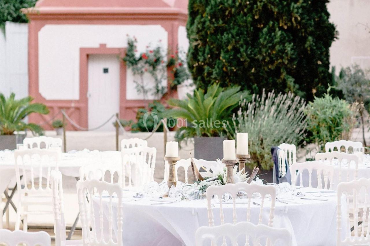 Tables rondes décorées pour un événement en extérieur, avec guirlandes lumineuses et végétation en arrière-plan.