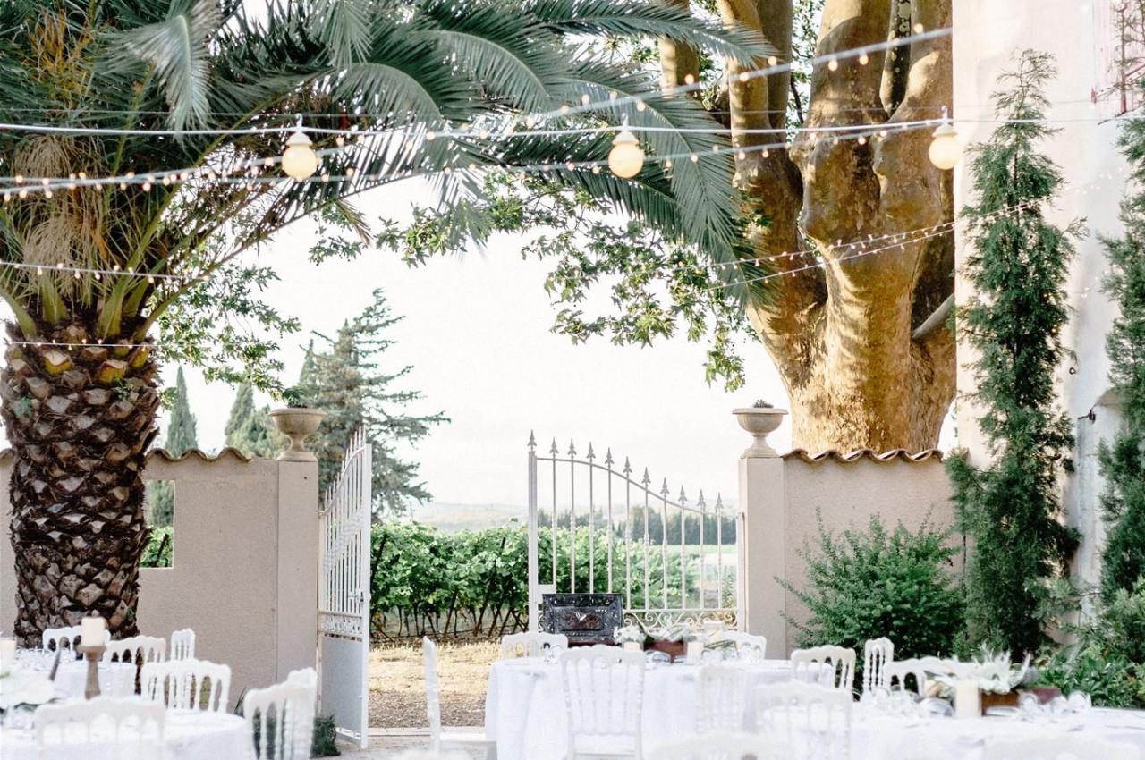 Tables blanches dressées en extérieur sous des guirlandes lumineuses, entourées de palmiers et d'arbres.