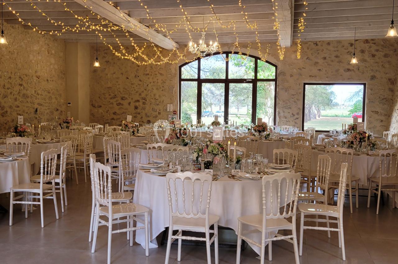 Salle de réception décorée avec des tables rondes, des chaises blanches et des guirlandes lumineuses suspendues.