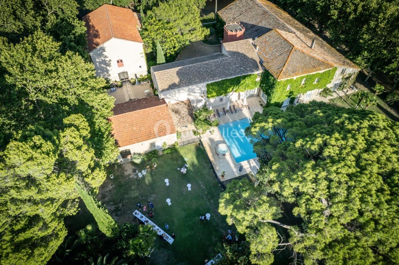 Vue aérienne d'une grande propriété avec piscine, bâtiments en tuiles et jardin arboré où des personnes sont rassemblées.