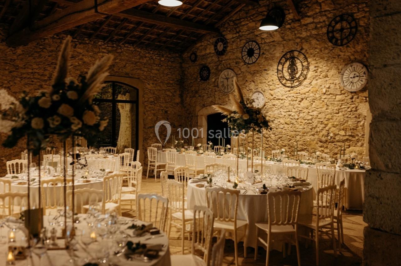 Salle de réception en pierre avec tables décorées, chaises blanches et horloges murales décoratives.