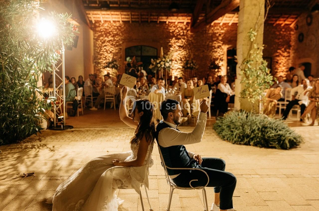 Un couple assis dos à dos lors d’un jeu de mariage, levant des pancartes dans une salle éclairée chaleureusement.