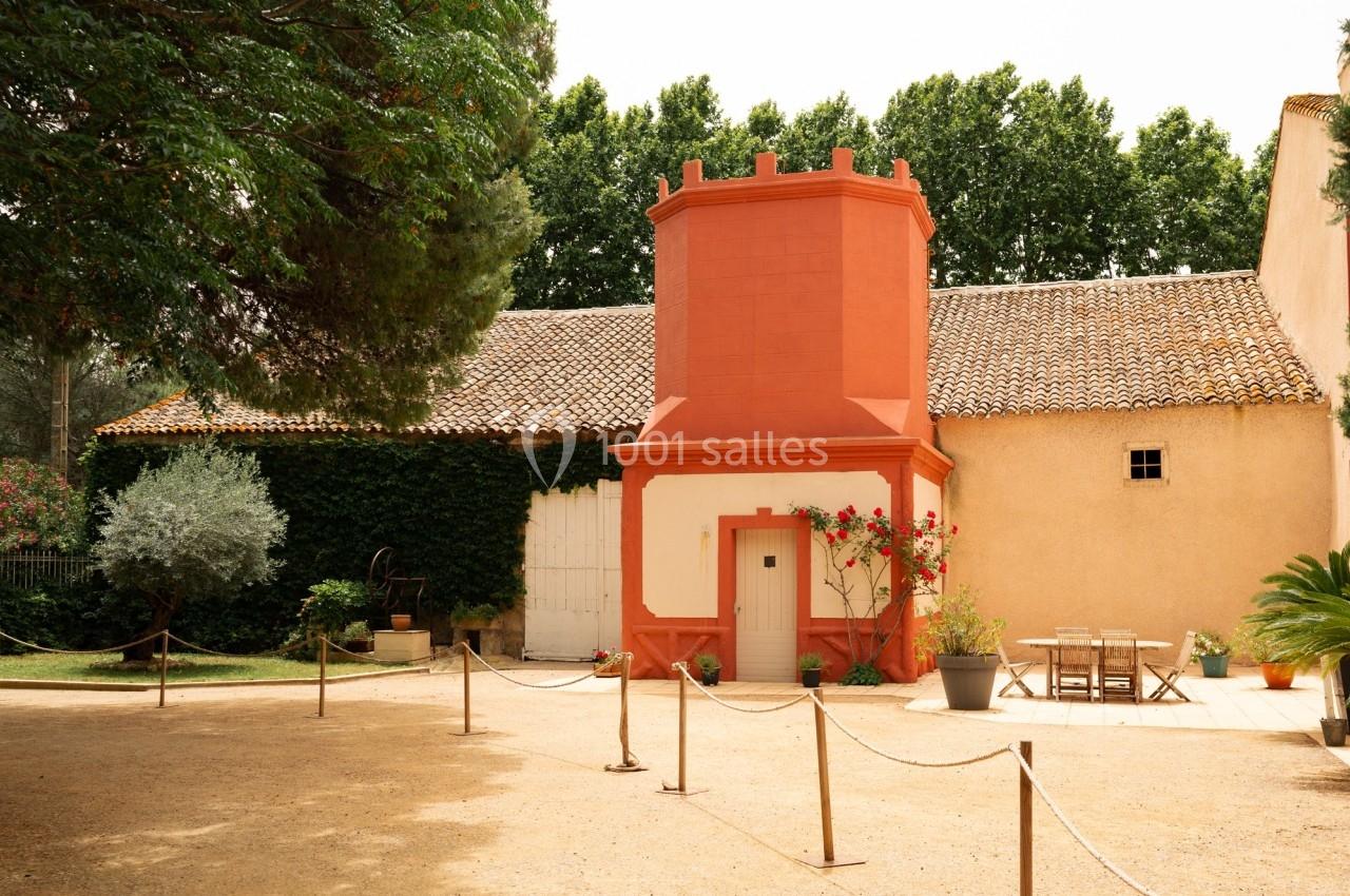 Bâtiment avec une tour rouge, entouré de végétation, une terrasse aménagée et une allée délimitée par des cordes.