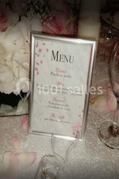 Menu encadré posé sur une table décorée de fleurs blanches et roses, avec un verre et une nappe texturée.