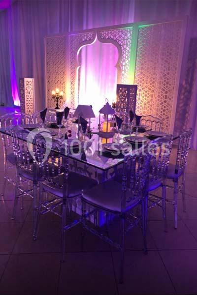 Table décorée avec des chaises transparentes, lanternes et bougies, devant un panneau ajouré éclairé de lumières colorées.