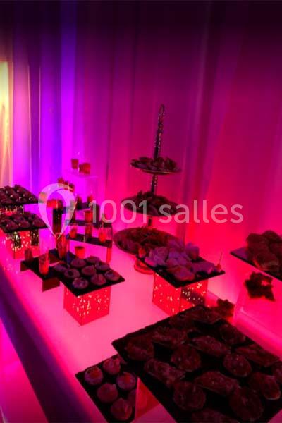 Table garnie de pâtisseries et de desserts variés, éclairée par une lumière rose et violette dans un décor festif.