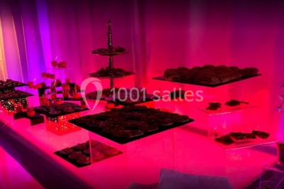 Table garnie de pâtisseries et de desserts variés, éclairée par une lumière rose et violette dans un décor festif.