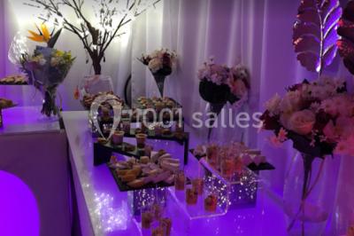Table garnie de pâtisseries et de desserts variés, éclairée par une lumière rose et violette dans un décor festif.