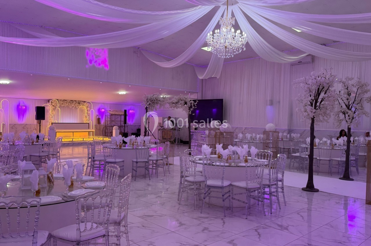 Salle de réception décorée avec des tables dressées, des chaises transparentes, des arbres blancs et un éclairage violet.