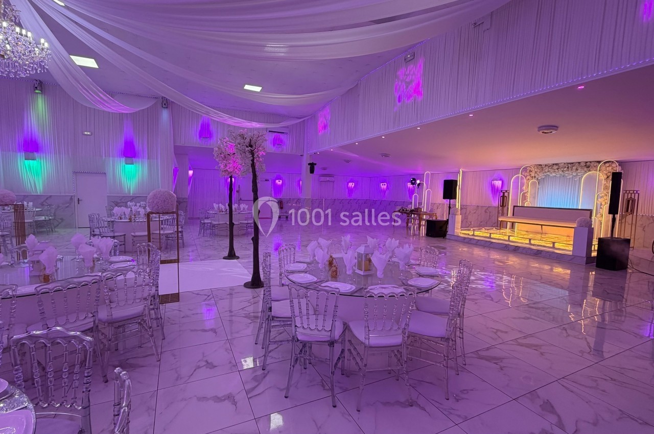 Salle de réception élégante avec tables dressées, éclairage violet, lustre et scène décorée.