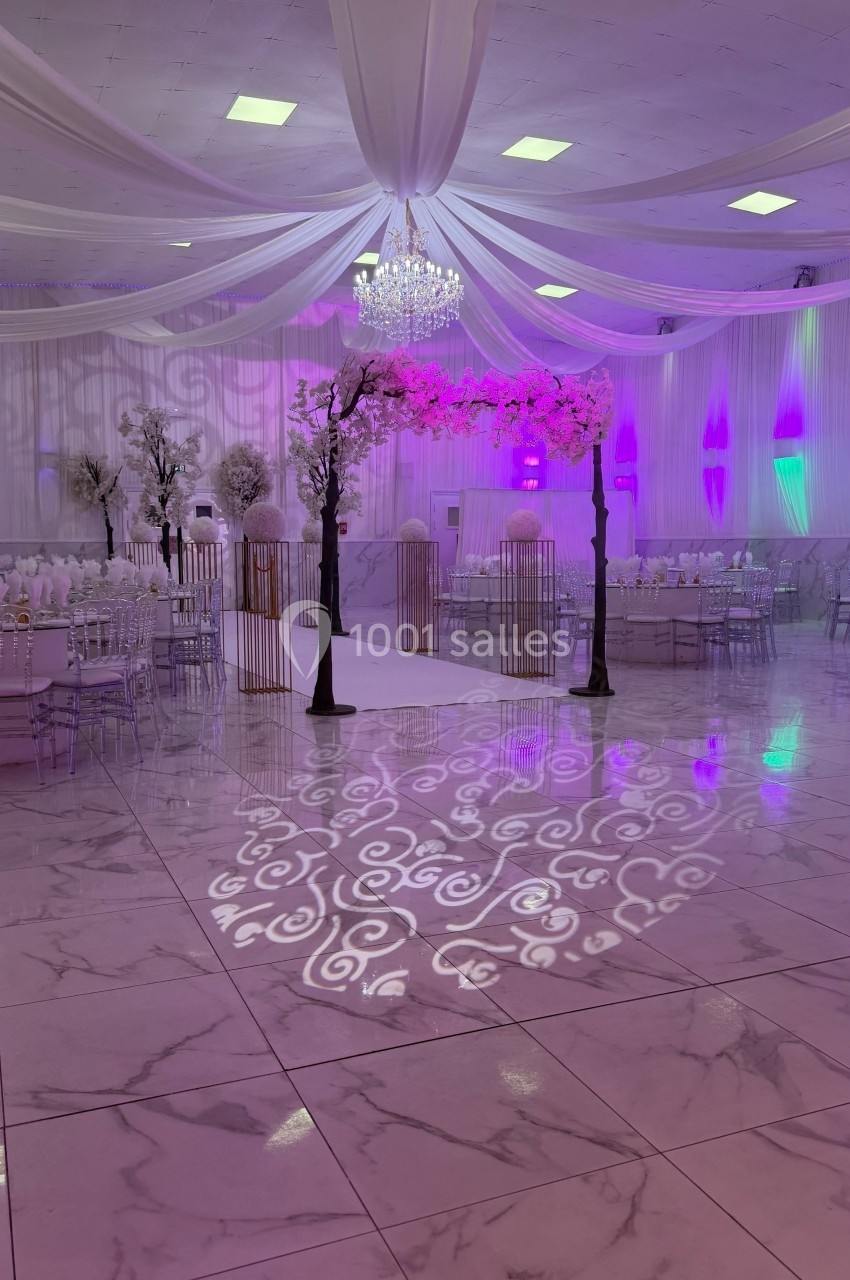 Salle de réception décorée avec des drapés blancs, éclairages violets, tables dressées et sol en marbre brillant.