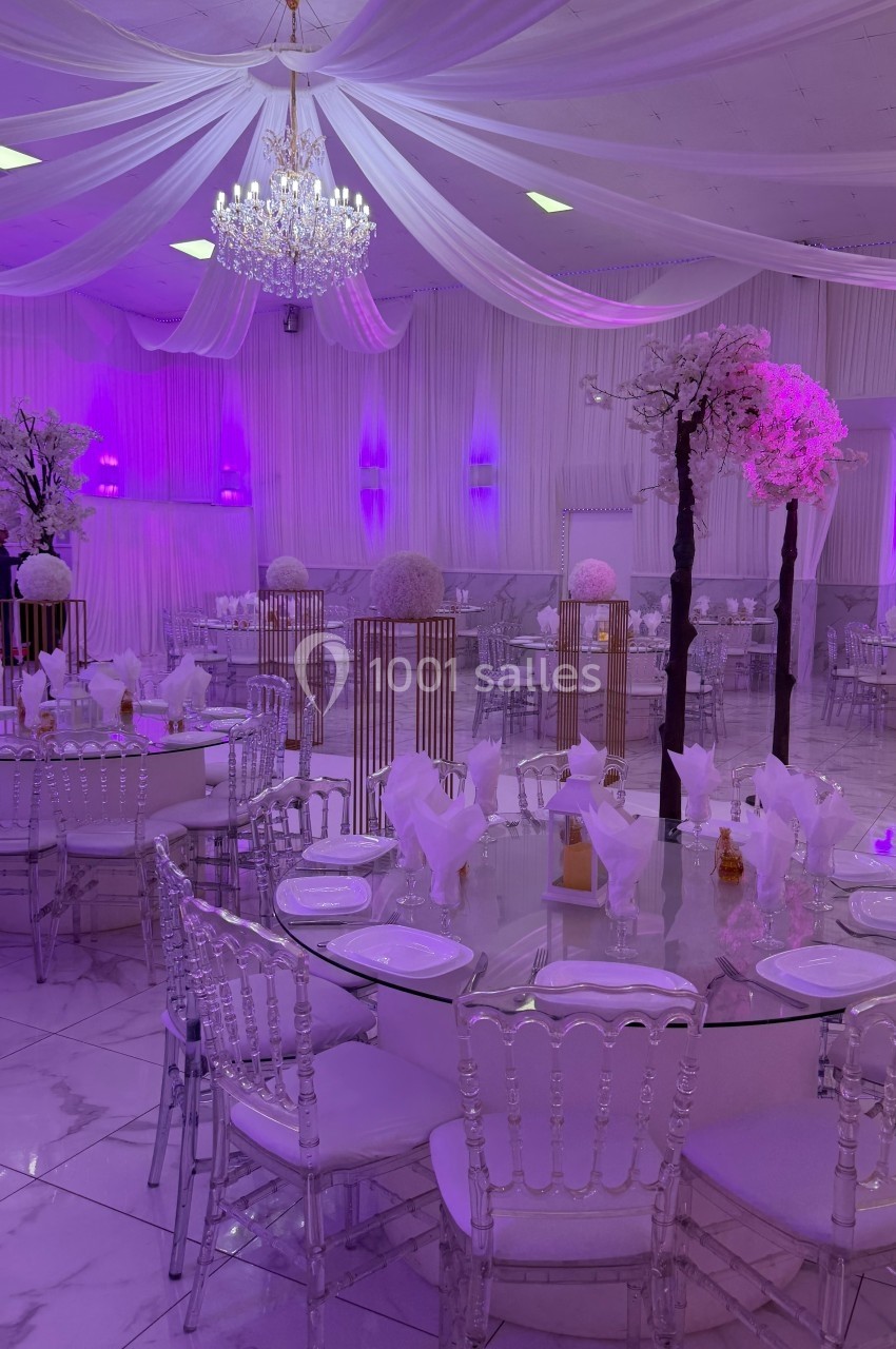 Salle de réception élégante avec tables rondes dressées, chaises transparentes et décoration lumineuse violette.