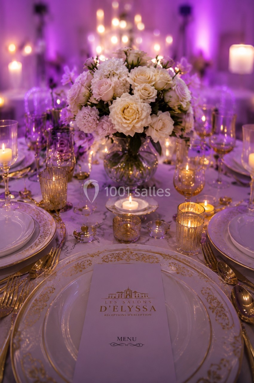 Table élégamment dressée avec vaisselle dorée, bougies allumées et bouquet central de fleurs blanches et roses.