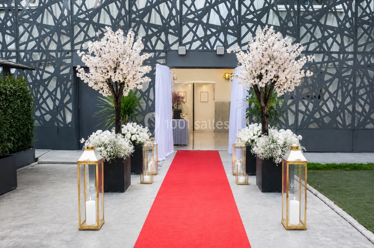 Entrée décorée avec tapis rouge, lanternes dorées, fleurs blanches et arbres fleuris devant un bâtiment moderne.