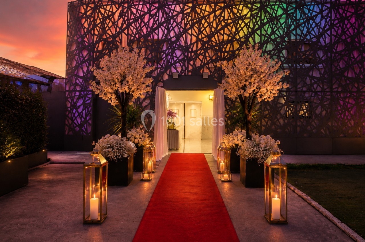Entrée décorée avec un tapis rouge, des lanternes allumées et des fleurs, devant un bâtiment illuminé de lumières colorées.