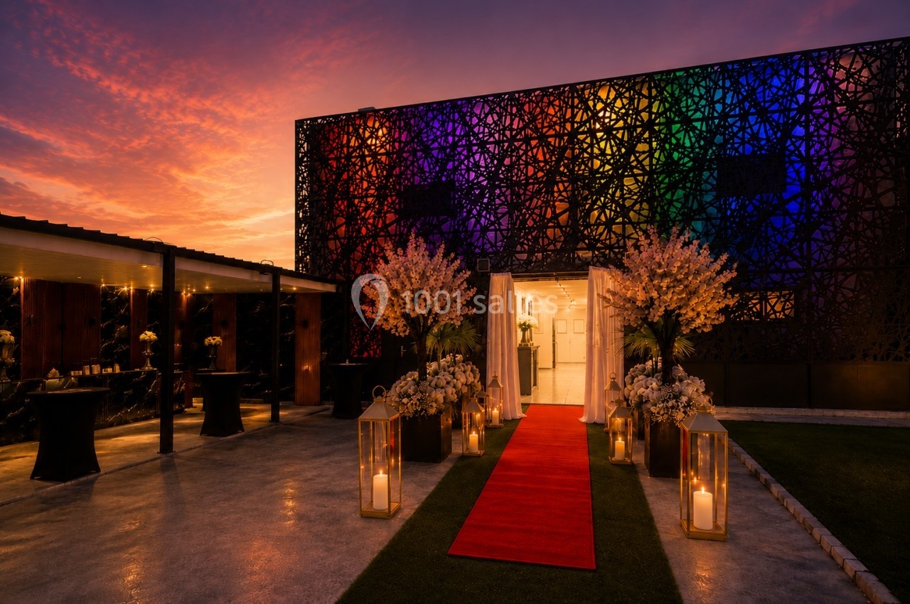 Entrée d'un bâtiment décorée pour un événement, avec tapis rouge, lanternes et éclairage coloré au coucher du soleil.