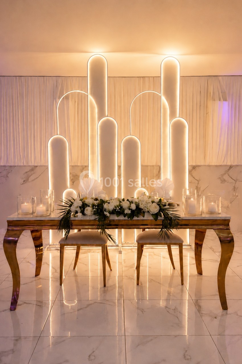 Table décorée avec des fleurs blanches et bougies, devant un fond lumineux avec des arches modernes.